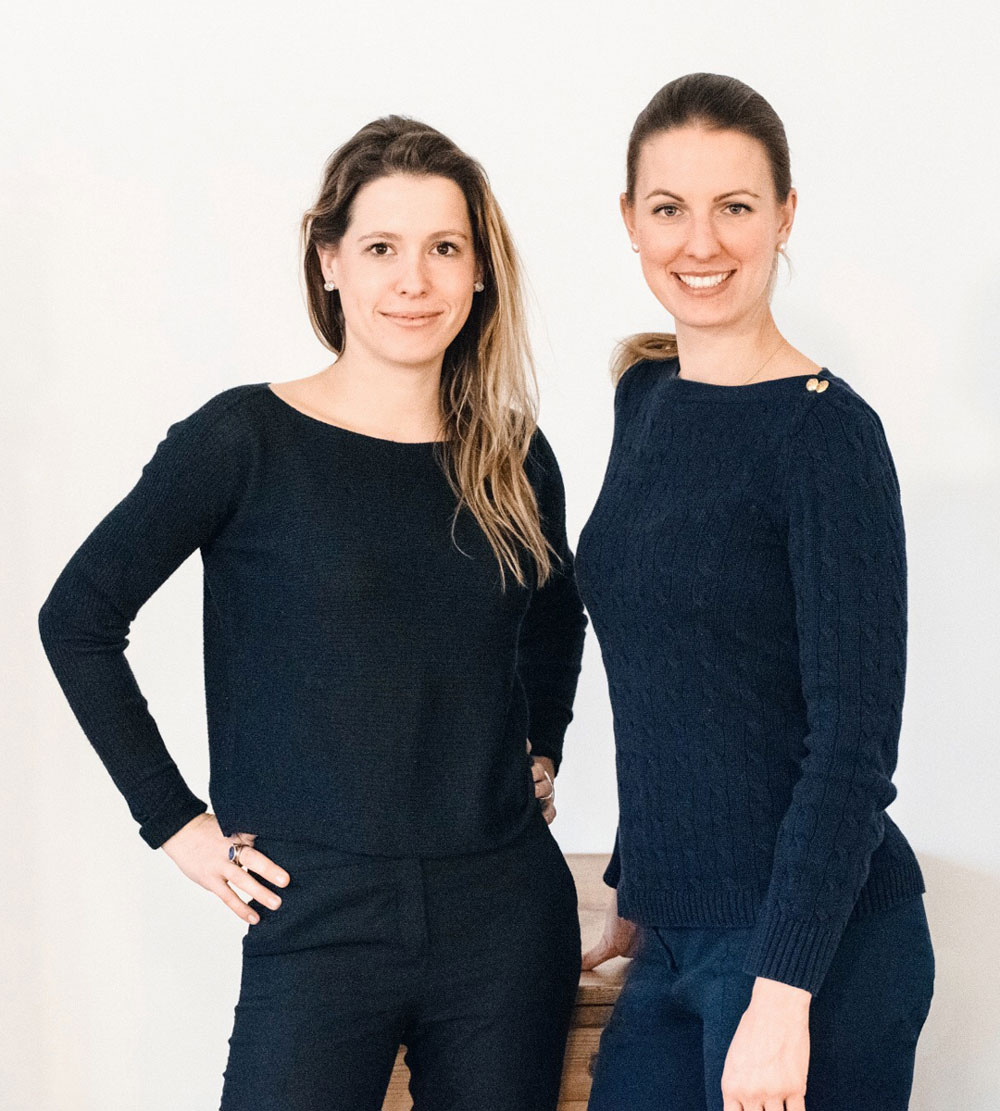 Zwei stehende schwarz gekleidete Frauen vor einer weißen Wand. Katharina Konrad und Anna Lina Schuhmacher - Art & Creative Director bei der DEBA GmbH.