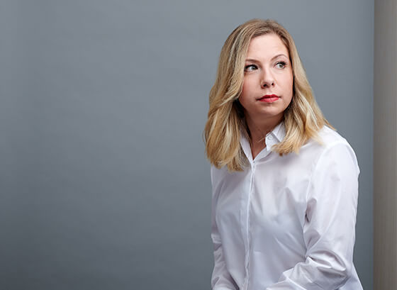 Portraitaufnahme einer blonden Frau in weißer Bluse vor grauem Hintergrund, die in die Ferne schaut. Zu sehen ist: Judith Plaßwilm, Corporate Communications Managerin DEBA.