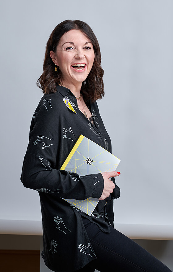 Foto einer lachenden dunkelhaarigen Frau, die ein Nozizbuch unter ihrem rechten Arm eingeklemmt hat. Zu sehen: Sabrina Hasselbusch, Corporate Development & Mindfulness Managerin DEBA.