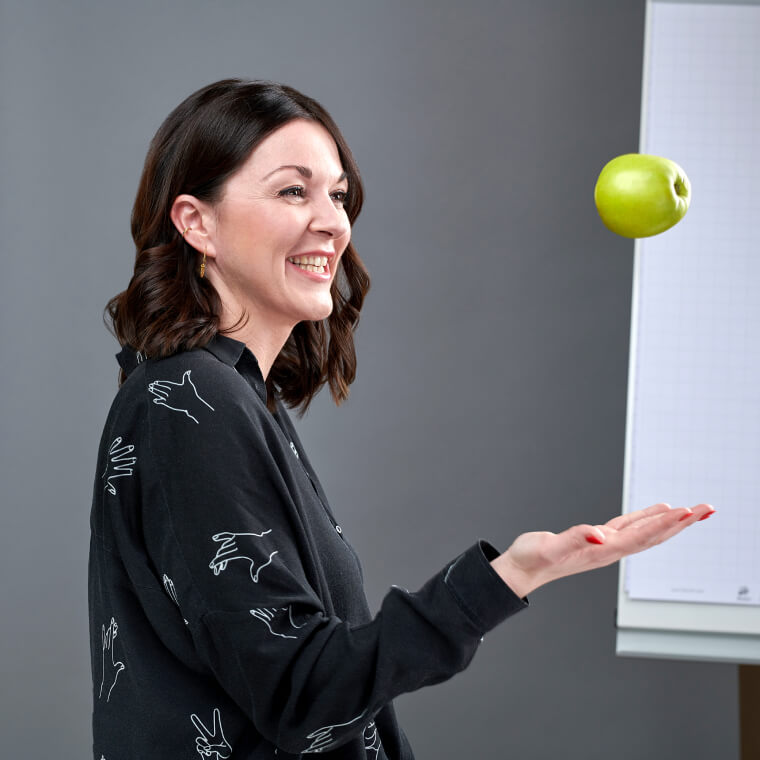 Foto einer dunkelhaarigen Frau, die lachend einen Apfel in die Luft wirft. Dabei handelt es sich um Sabrina Hasselbusch, Corporate Development & Mindfulness Managerin DEBA.