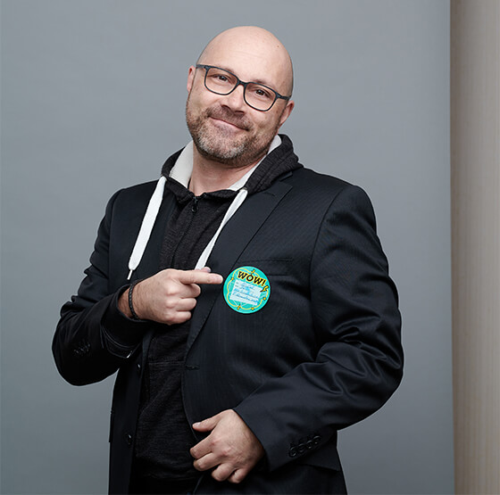 Foto von einem glatzköpfigen Mann mit Brill, der mit seinem Zeigefinger auf einen türkisfarbenen Sticker auf seiner Brust zeigt. Bei dem Mann handelt es sich um Michel Ganouchi, Lead Schweiz DEBA.