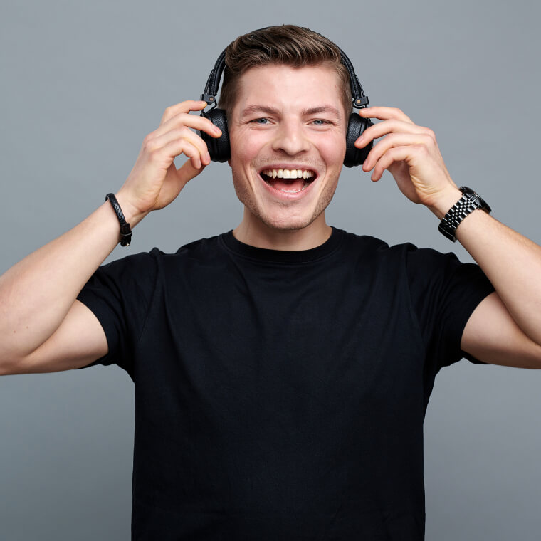 Foto von einem lachenden jungen Mann in einem schwarzen T-Shirt mit Kopfhörer auf den Ohren. Seine Hände liegen auf dem Kopfhörer. Der Hintergrund ist grau. Zu sehen: Leonard Heygster, Project Manager DEBA.