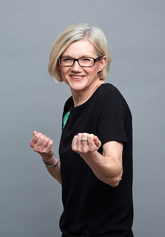 Foto einer weißblonden Frau mit Brille in dunklem Oberteil von einem grauen Hintergrund. Die Frau macht eine Power Pose. Zu sehen: Susanne Siebrecht, Associated Consultant DEBA.