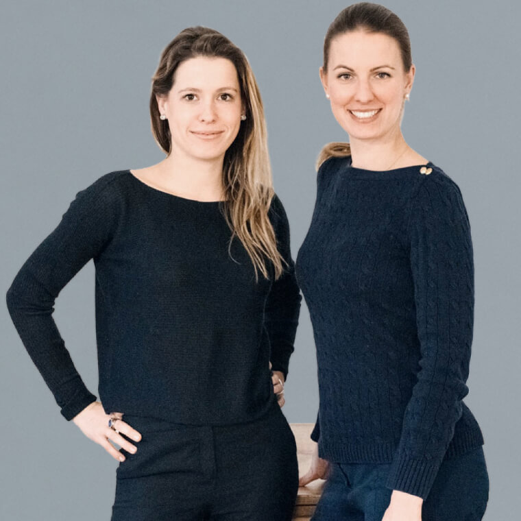 Aufnahme von zwei stehenden dunkel gekleideten Frauen vor grauem Hintergrund. Zu sehen sind: Katharina Konrad & Anna Lina Schuhmacher, beide Creative Art Director der DEBA GmbH.