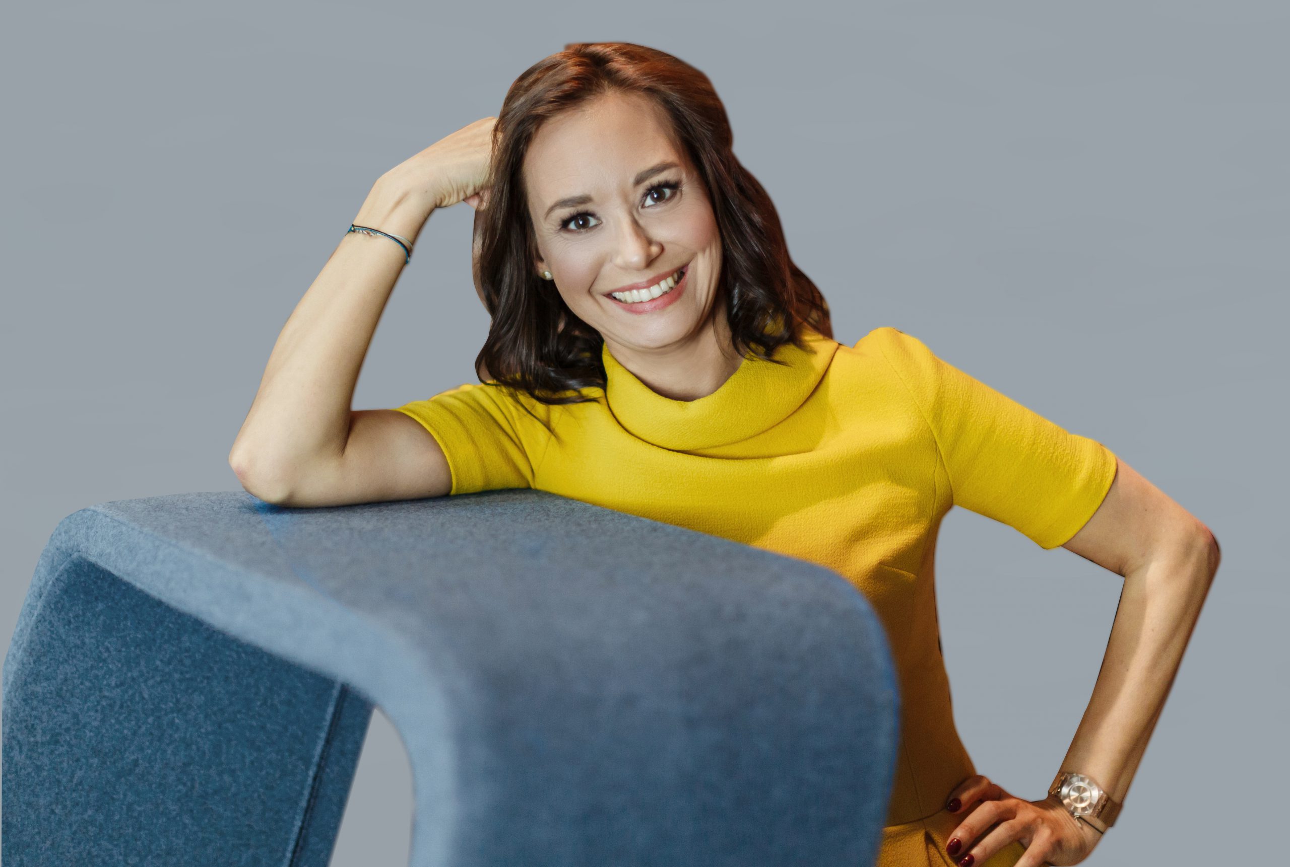 Foto einer lächelnden brünetten Frau, die sich mit ihrem rechten Arm auf einem Desk abstützt. Sie tägt ein gelbes Kleid. Dabei handelt es sich um Alina Ting, Associated Consultant DEBA.